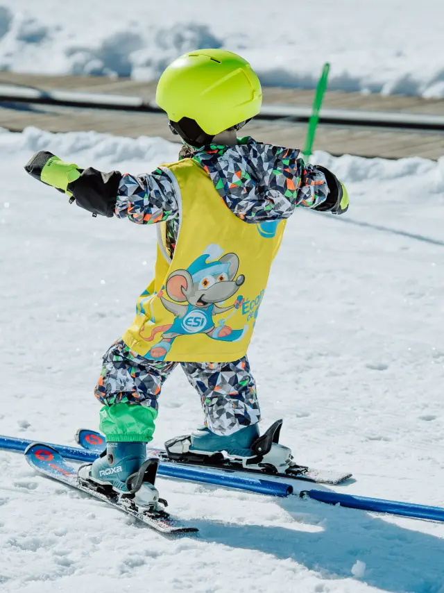 Enfant en combinaison de ski jaune fluo avec motifs de souris, équipé de skis et d'un casque évoluant dans le jardin d'enfants ESI