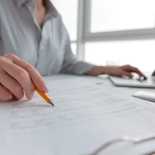 Close up portrait of a woman holding pencil over paper documents while sitting and working with laptop computer at home