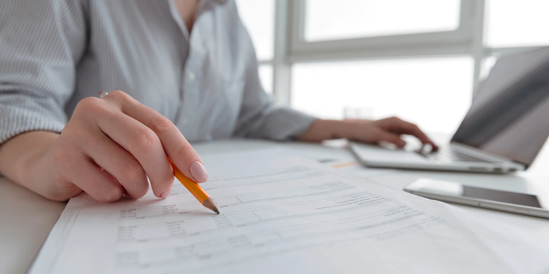 Close up portrait of a woman holding pencil over paper documents while sitting and working with laptop computer at home