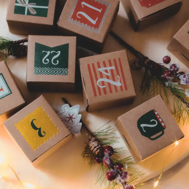 Craft advent calendar box with numbers, inscriptions and Christmas decoration with a burning garland on a wooden tray lie on the bed in the bedroom, close-up top view.
