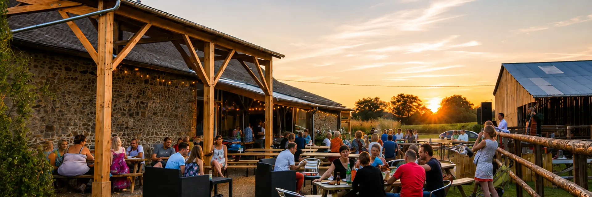 Plusieurs personnes sont réunies autour de tables en plein air, dans un cadre naturel bordé de granges. Des guirlandes lumineuses suspendues au-dessus d’eux illuminent la soirée alors qu’ils partagent un moment convivial au coucher du soleil