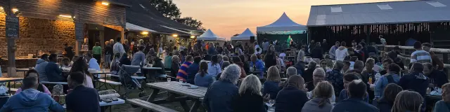 Des visiteurs installés à de grandes tables en bois profitent d'une soirée festive en plein air devant des bâtiments de ferme et une scène au coucher du soleil.