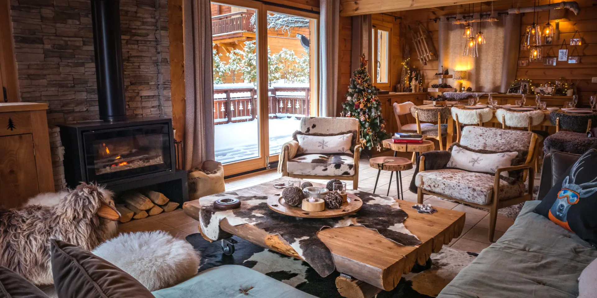 Intérieur d’un chalet en bois avec cheminée allumée, mobilier chaleureux et vue sur l’extérieur enneigé.