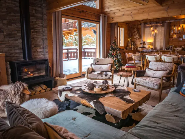 Intérieur d’un chalet en bois avec cheminée allumée, mobilier chaleureux et vue sur l’extérieur enneigé.