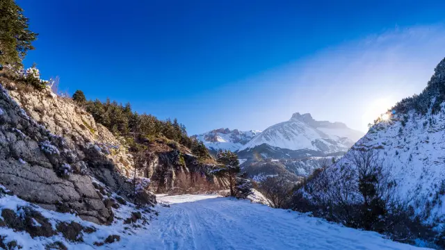 Besneeuwd pad tussen rotswanden en dennenbomen boven La Cluse, met de toppen van het Dévoluy-massief op de achtergrond onder een blauwe lucht.