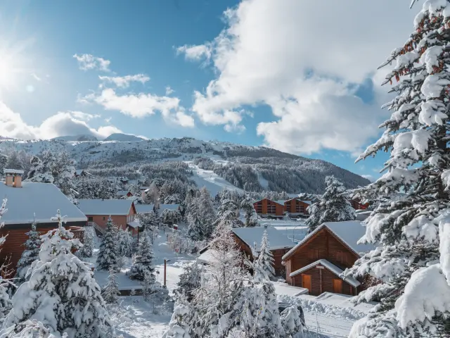Des chalets enneigés à La Joue du Loup avec les pistes de ski en arrière-plan, dans le Dévoluy, Hautes-Alpes.