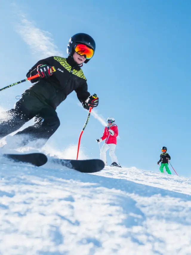 Quatre skieurs, dont des enfants, descendent une piste enneigée sous un ciel parfaitement bleu.
