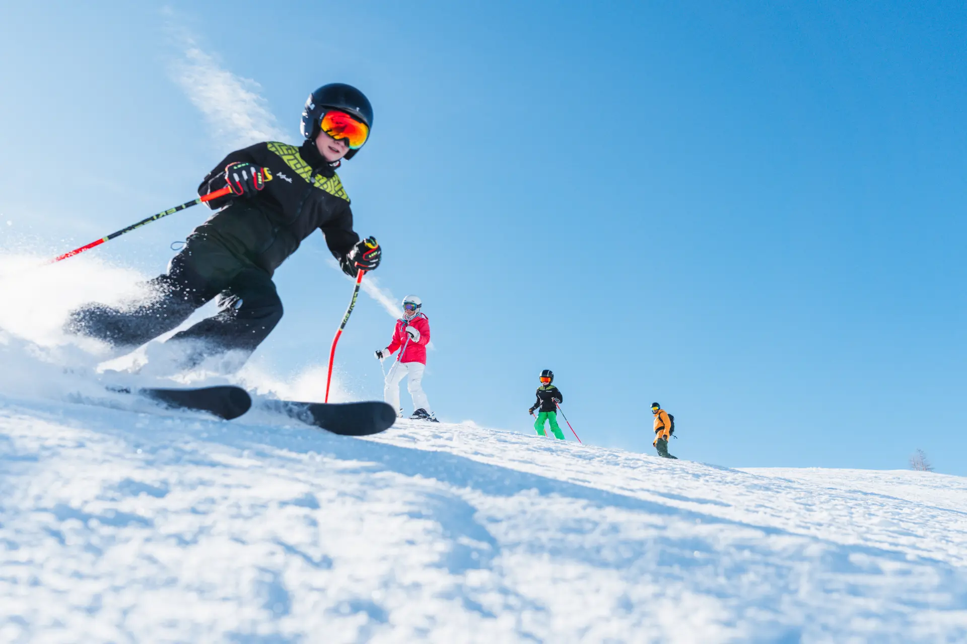 Four skiers, including children, skiing down a snowy slope under a clear blue sky.