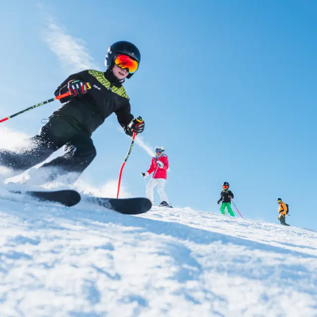 Vier skiërs, waaronder kinderen, dalen een besneeuwde piste af onder een heldere blauwe lucht.