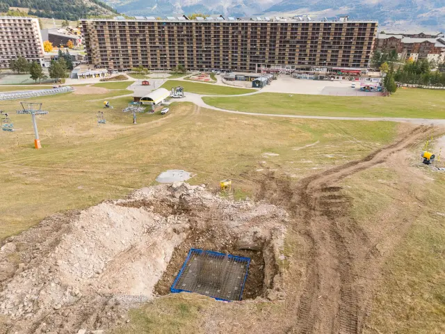 Vue aérienne d’une excavation au front de neige avec ferraillage prêt pour une fondation en béton, bâtiments de la station en arrière-plan.