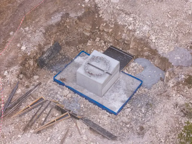 Vue aérienne rapprochée d’un massif en béton avec platine métallique, au fond d’une excavation rocheuse pour un pylône de télécabine.