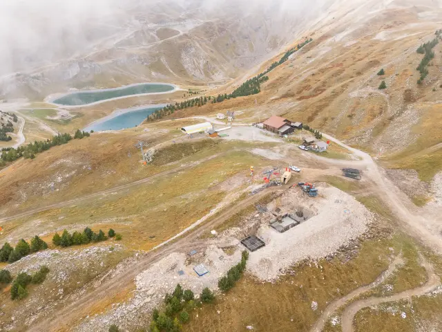 Vue aérienne d’un chantier en montagne avec excavation, engins et bâtiments techniques près de retenues d’eau.