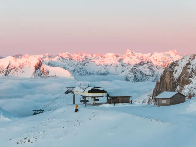 Summit of the Dévoluy ski area in Sommarel, with a ski lift surrounded by snow and a sea of clouds facing the Alpine peaks at sunrise.