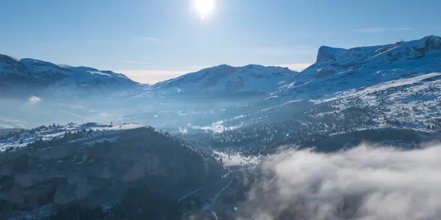Paysage hivernal du Dévoluy avec montagnes enneigées, vallée embrumée et soleil haut dans le ciel.