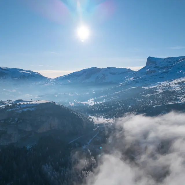 Paysage hivernal du Dévoluy avec montagnes enneigées, vallée embrumée et soleil haut dans le ciel.