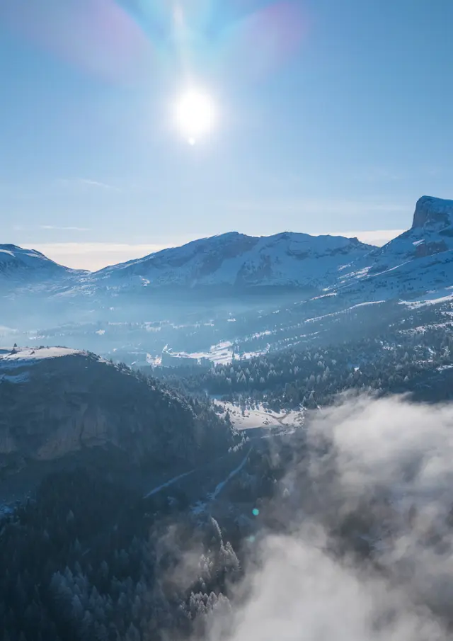 Paysage hivernal du Dévoluy avec montagnes enneigées, vallée embrumée et soleil haut dans le ciel.