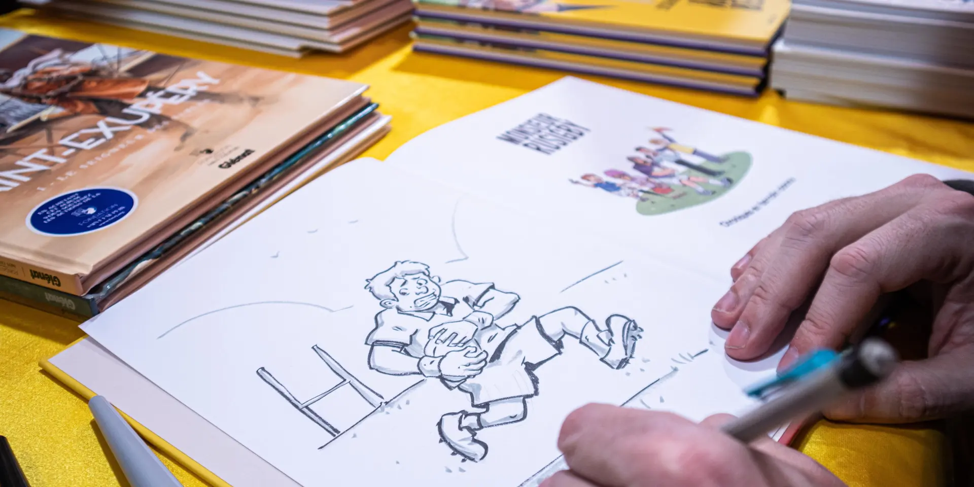 Een illustrator tekent een stripverhaal aan een tafel, omringd door stripboeken tijdens Les Givrés de la BD.