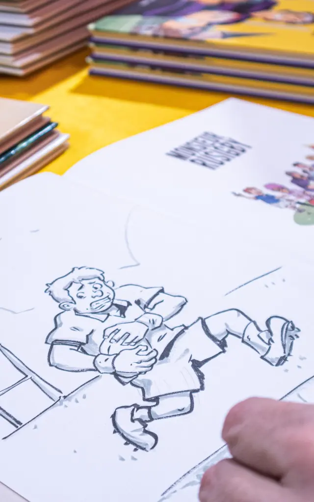 Un illustrateur dessine une bande dessinée sur une table, entouré d’albums lors de l’événement Les Givrés de la BD.