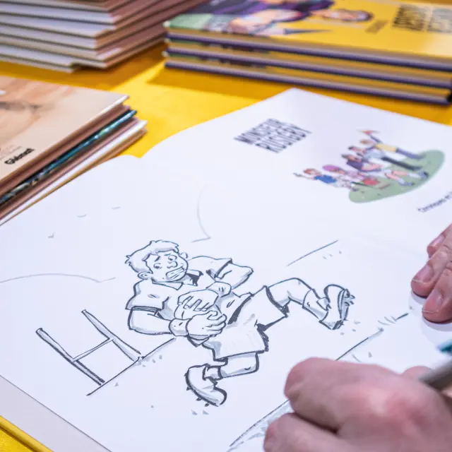 Een illustrator tekent een stripverhaal aan een tafel, omringd door stripboeken tijdens Les Givrés de la BD.
