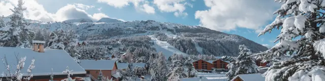 Chalets en bois entourés deufs d’arbres enneigés à La Joue du Loup avec les pistes en arrière-plan.