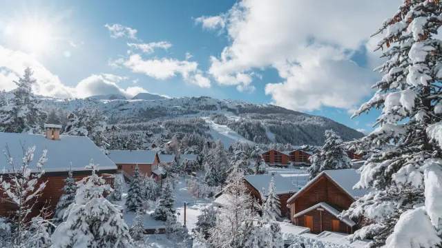 Wooden chalets surrounded by snow-covered trees in La Joue du Loup with ski slopes in the background.