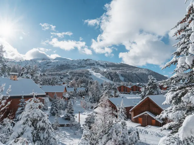 Wooden chalets surrounded by snow-covered trees in La Joue du Loup with ski slopes in the background.