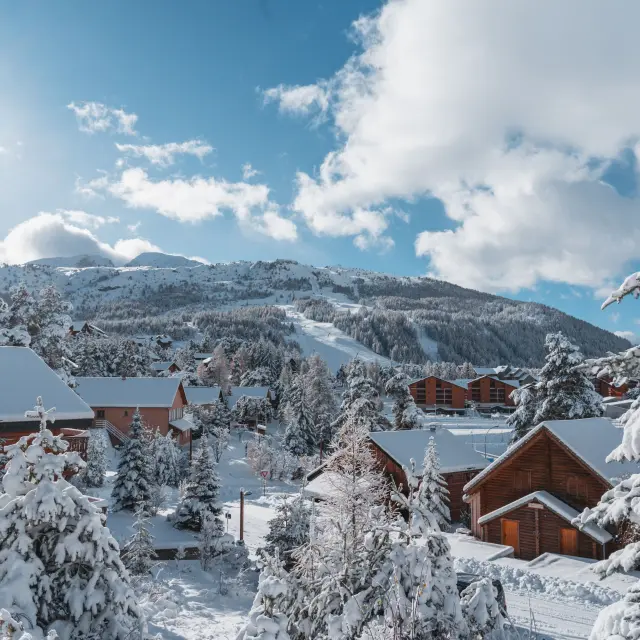 Chalets en bois entourés deufs d’arbres enneigés à La Joue du Loup avec les pistes en arrière-plan.