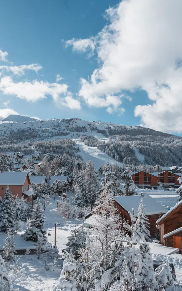 Chalets en bois entourés deufs d’arbres enneigés à La Joue du Loup avec les pistes en arrière-plan.