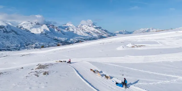 Two dog sled teams move along a snowy trail with the mountains of Le Dévoluy in the background.
