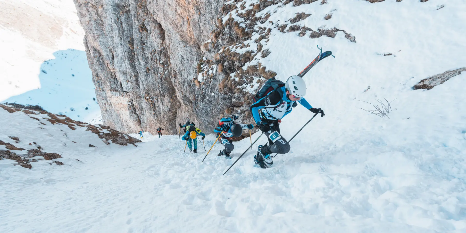 Ski-alpinisten klimmen te voet een steile besneeuwde helling op langs een rotswand.