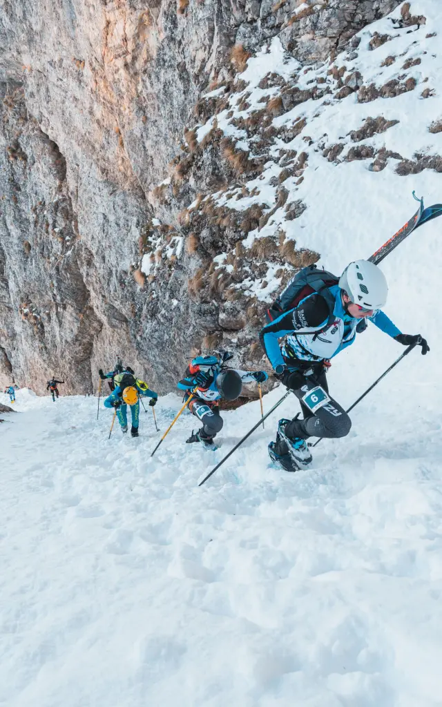Ski mountaineers climb a steep snowy slope at the base of a rocky cliff.