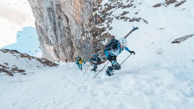 Des skieurs-alpinistes progressent à pied dans une pente raide et enneigée au pied d’une paroi rocheuse.