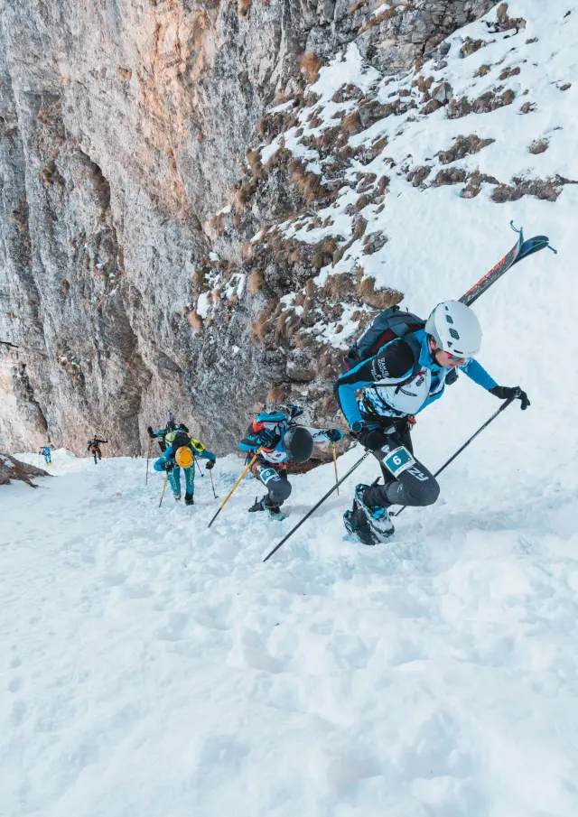 Ski mountaineers climb a steep snowy slope at the base of a rocky cliff.