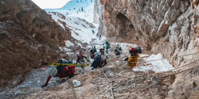 Ski-alpinisten klimmen aangelijnd door een steile rotsachtige en besneeuwde passage met vaste touwen.
