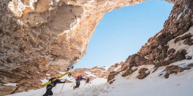 Ski-alpinisten lopen onder een natuurlijke rotsboog door een steile besneeuwde doorgang.