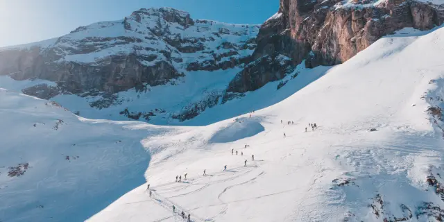 Een lange rij ski-alpinisten trekt over een besneeuwde helling onder hoge rotswanden.