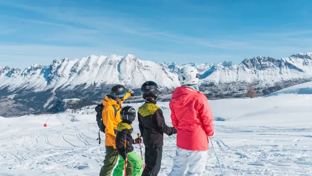Two adults and two children on skis կանգ stopped on a snowy slope, looking at the peaks of the Dévoluy massif under a clear blue sky.