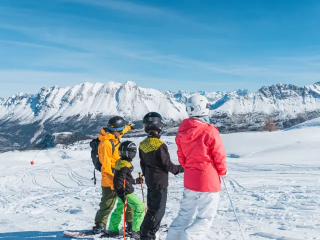 Deux adultes et deux enfants à ski, arrêtés sur une piste enneigée, regardant les sommets du massif du Dévoluy sous un ciel bleu dégagé.
