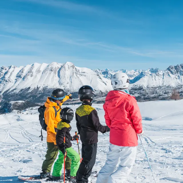 Twee volwassenen en twee kinderen op ski’s staan stil op een besneeuwde piste en kijken naar de toppen van het Dévoluy-massief onder een heldere blauwe lucht.