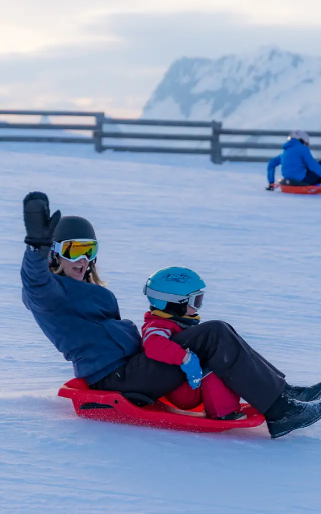 Een volwassene en een kind zitten op een rode slee en glijden naar beneden op een besneeuwde helling, met op de achtergrond iemand anders op een slee en bergen in de verte.