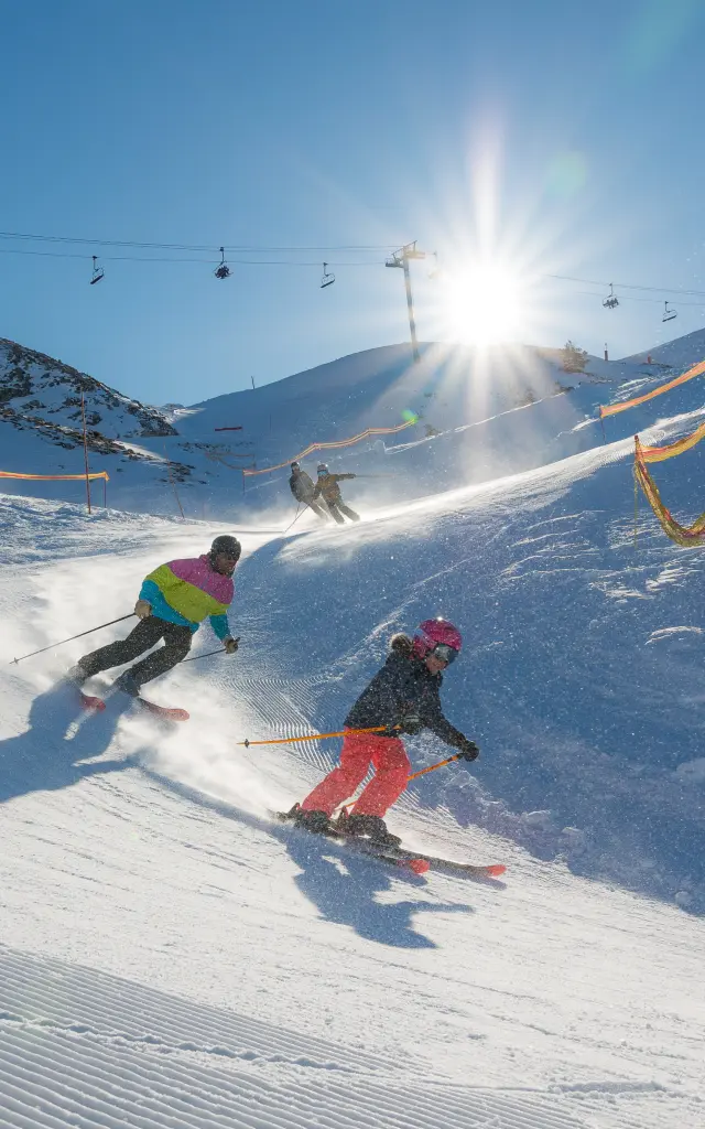 Een volwassene en een kind skiën naar beneden op een geprepareerde piste en maken een bocht, met de zon voor hen en skiliften op de achtergrond.