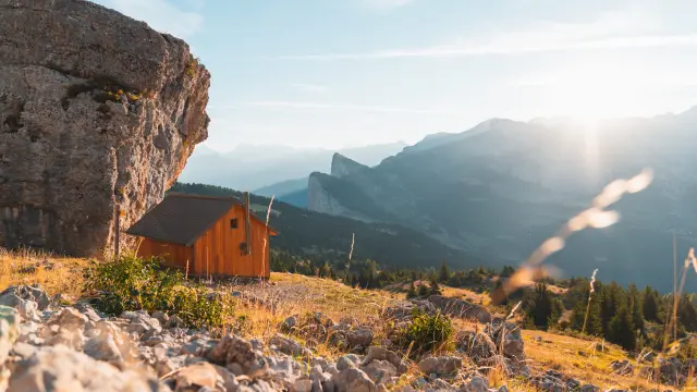Petite cabane en bois au pied d’une falaise, dominant un paysage de montagnes et de forêts au soleil.