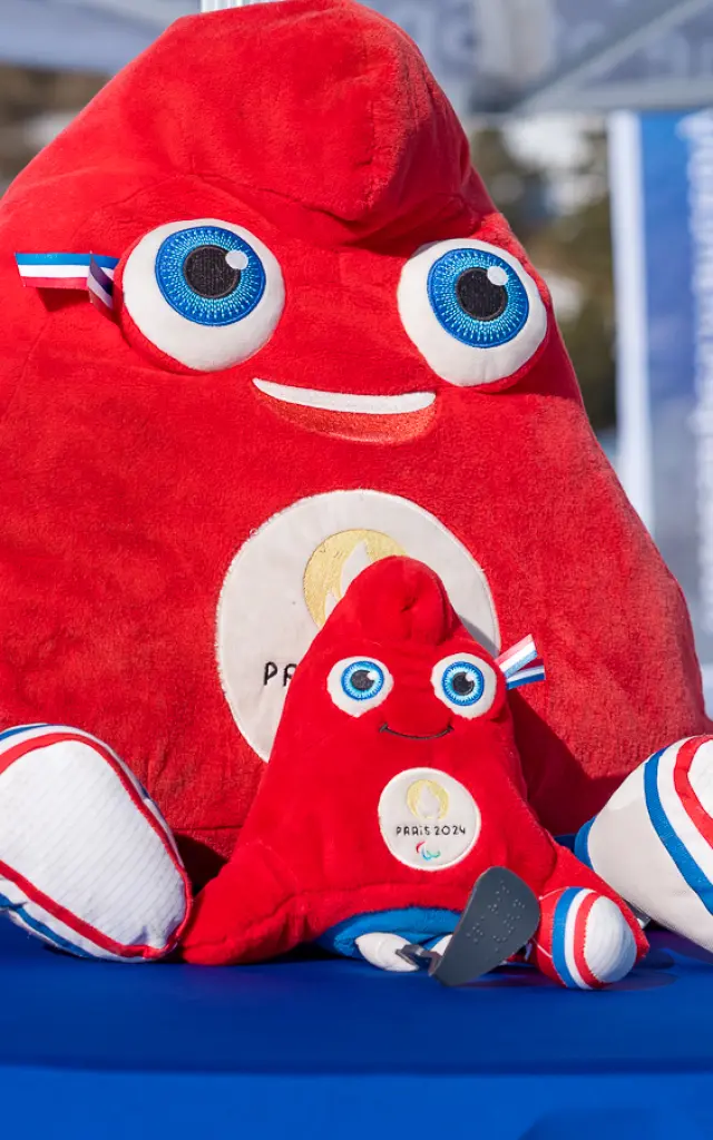 Deux peluches de la mascotte rouge Paris 2024 posées sur une table en extérieur dans une station de ski.