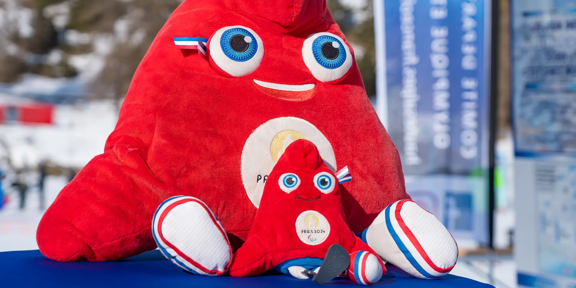 Deux peluches de la mascotte rouge Paris 2024 posées sur une table en extérieur dans une station de ski.