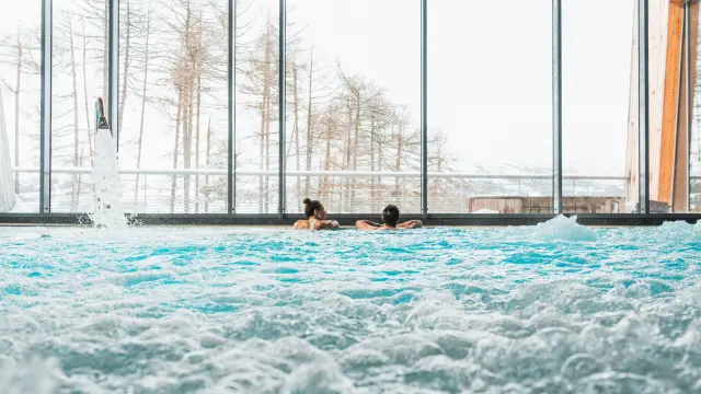 Two people relaxing in a bubbling indoor pool in front of large windows overlooking snowy scenery.