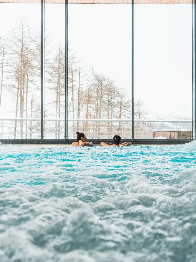 Two people relaxing in a bubbling indoor pool in front of large windows overlooking snowy scenery.