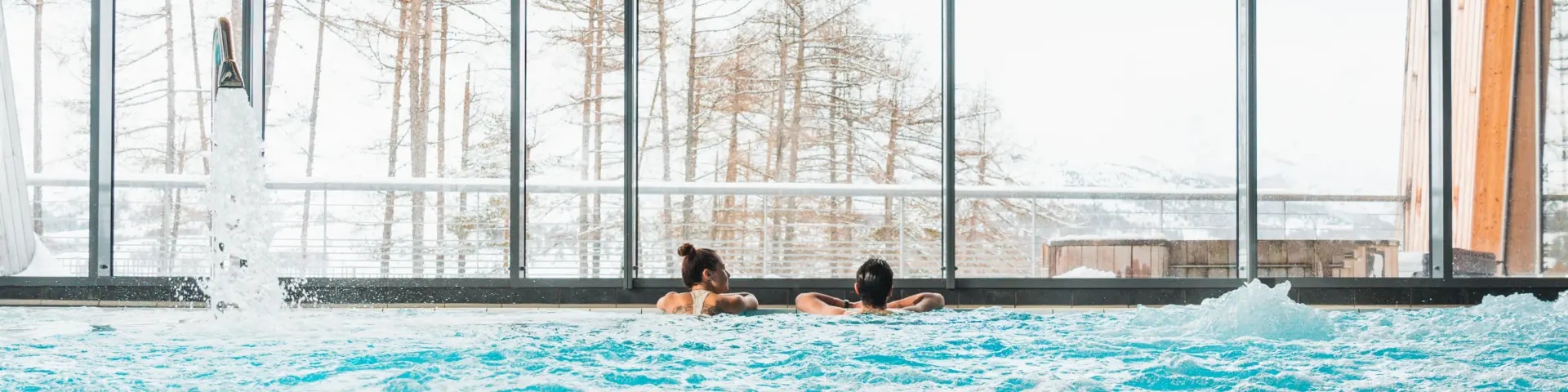 Deux personnes se relaxent dans une piscine intérieure à bulles devant de grandes baies vitrées ouvertes sur un décor enneigé.