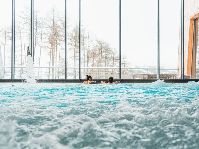 Two people relaxing in a bubbling indoor pool in front of large windows overlooking snowy scenery.