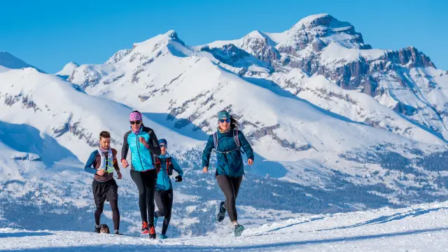 A group of runners trail running on a snowy mountain path with high peaks in the background.