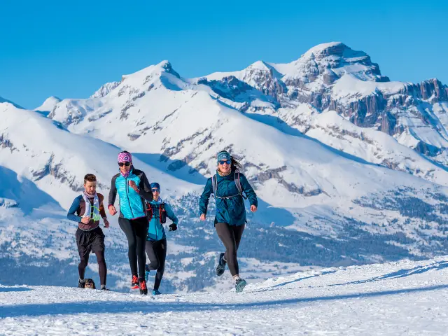 Een groep hardlopers loopt over een besneeuwd bergpad met hoge bergtoppen op de achtergrond.